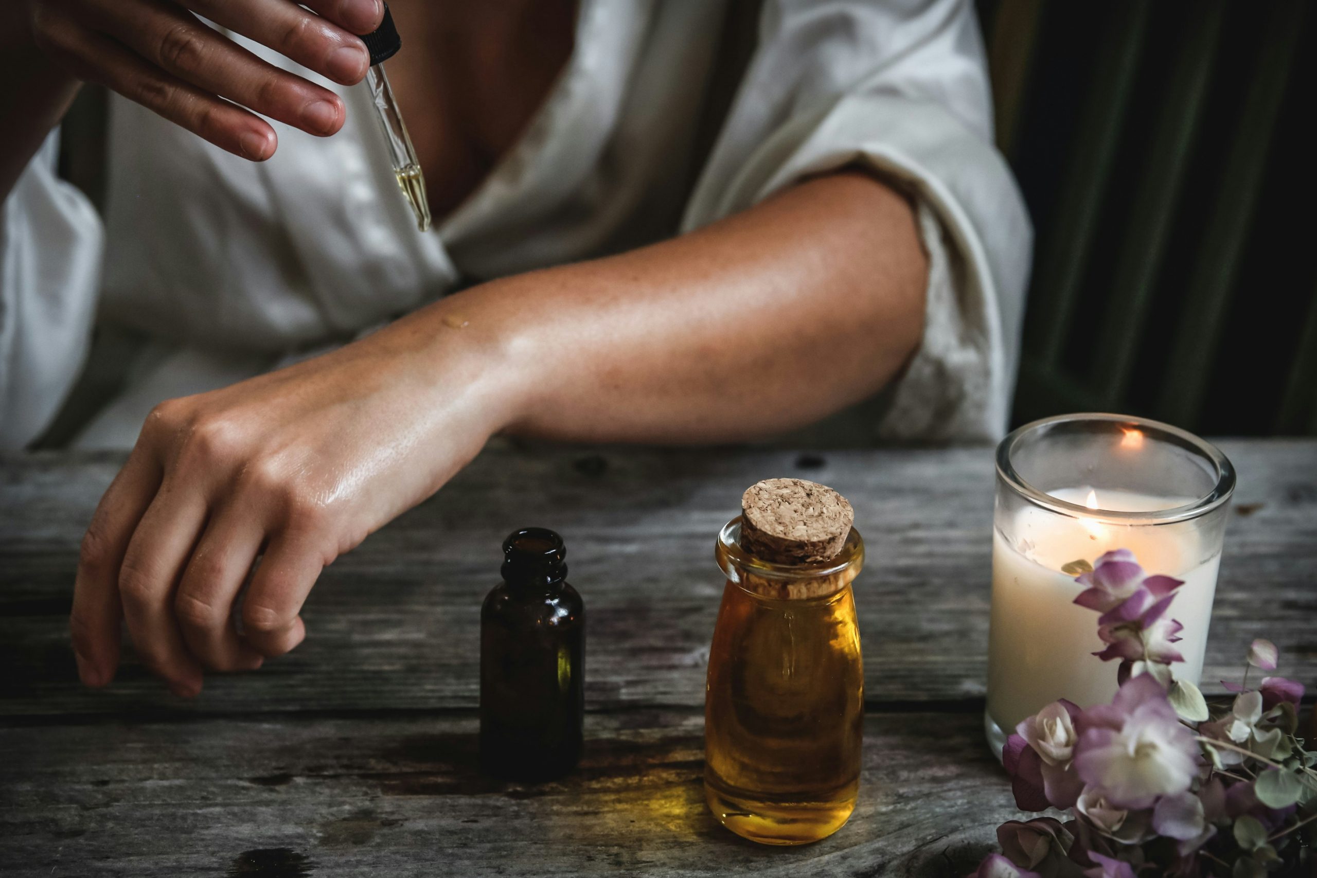 A darkly lit image of a woman dropping oil on her arm with a pipette. Some glass jars of oil sit on the table in front of her along with a lit candle.