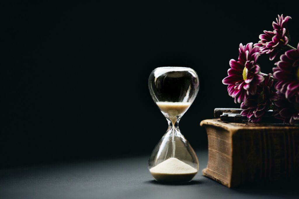 A dark image of an hourglass with sand pouring through it. There is a book and flower laid nearby.